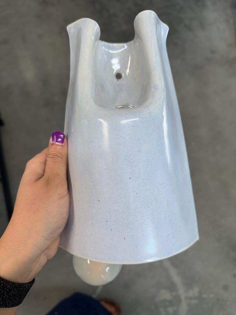 K&ouml;nnte beinhalten: Ein hellblauer Wandpflanztopf aus Keramik mit einer glatten, gesprenkelten Oberfl&auml;che. Das Gef&auml;&szlig; verf&uuml;gt &uuml;ber eine breite, ausgestellte &Ouml;ffnung an der Oberseite mit einem kleinen kreisf&ouml;rmigen Loch zur Befestigung und einen abgerundeten Boden.