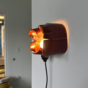 May include: A unique, dark red wall sconce with a distinctive, folded design. The sconce features an exposed light bulb, casting a warm, orange glow. A black cord extends from the base, connecting to a power source. The fixture is mounted on a white wall.
