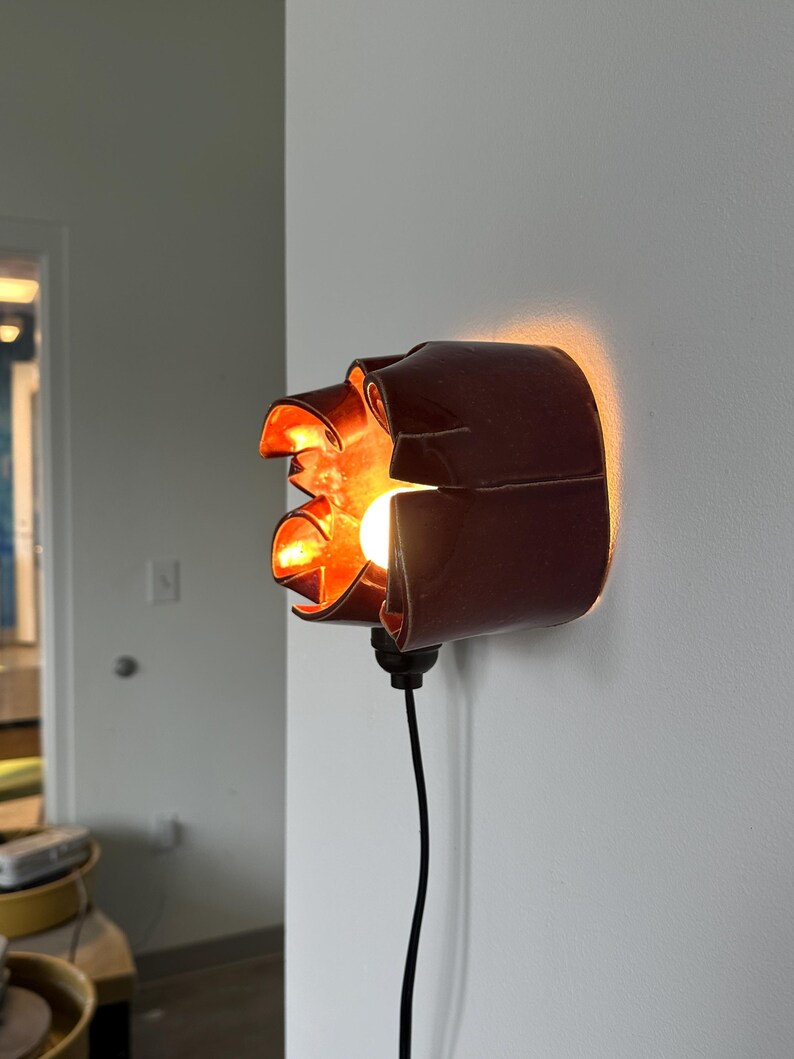 May include: A unique, dark red wall sconce with a distinctive, folded design. The sconce features an exposed light bulb, casting a warm, orange glow. A black cord extends from the base, connecting to a power source. The fixture is mounted on a white wall.