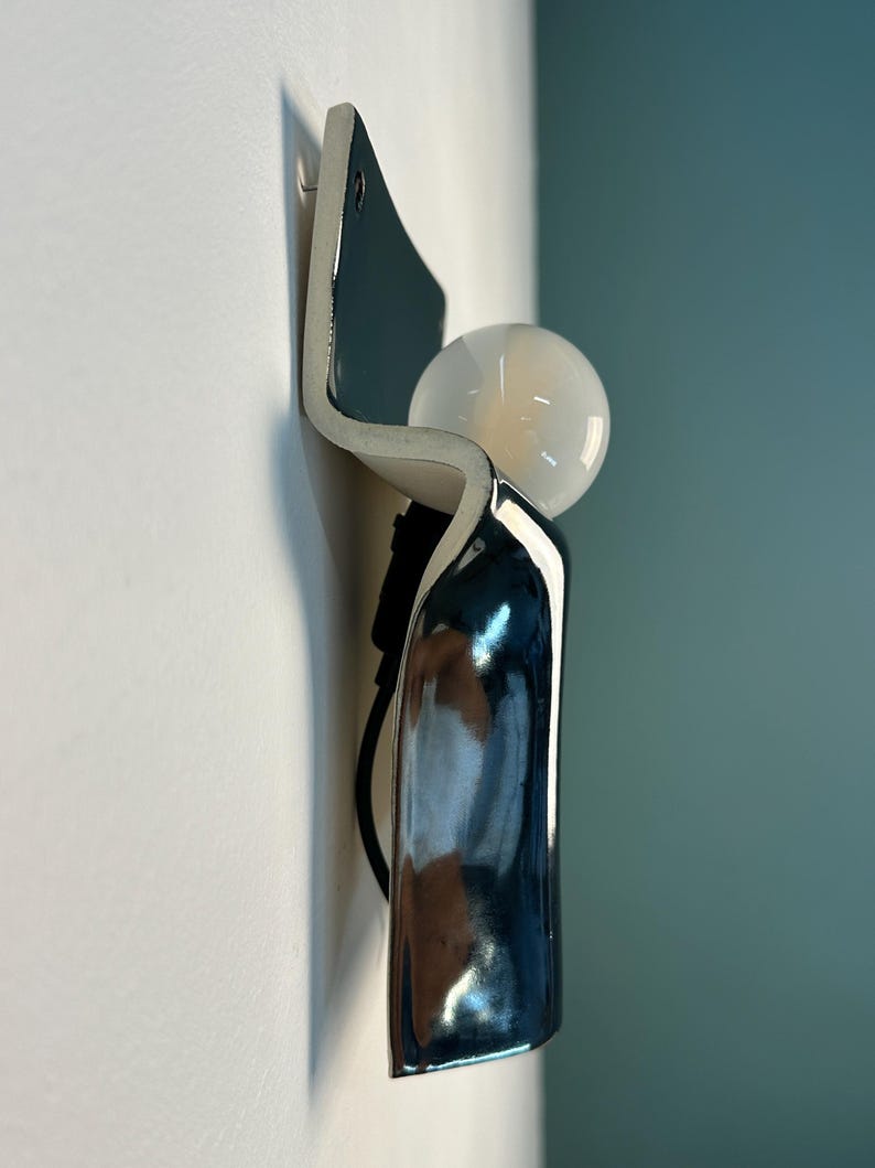 Peut inclure: Une applique murale compos&eacute;e d'un corps incurv&eacute; en c&eacute;ramique ou en m&eacute;tal bleu fonc&eacute; brillant avec une finition lisse et r&eacute;fl&eacute;chissante. Une ampoule sph&eacute;rique en verre blanc est plac&eacute;e dans la courbe sup&eacute;rieure du luminaire, vers l'ext&eacute;rieur.