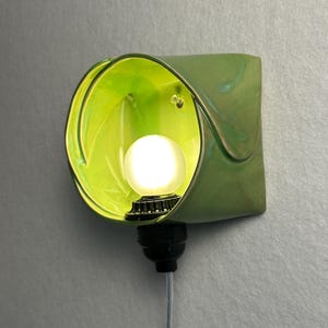 May include: A wall-mounted lamp featuring a curved, lime green ceramic shade with a glossy finish. A small, round, frosted white light bulb is centered within the shade, attached to a black socket base with a thin, light-colored electrical cord.