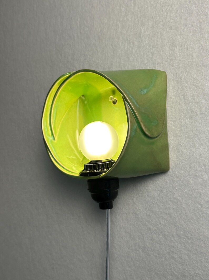 May include: A wall-mounted lamp featuring a curved, lime green ceramic shade with a glossy finish. A small, round, frosted white light bulb is centered within the shade, attached to a black socket base with a thin, light-colored electrical cord.