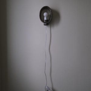 May include: A wall-mounted light fixture with a bulb inside a dark, oval-shaped metal frame. The fixture is connected to a power outlet via a thin, silver cord. The light is turned on.