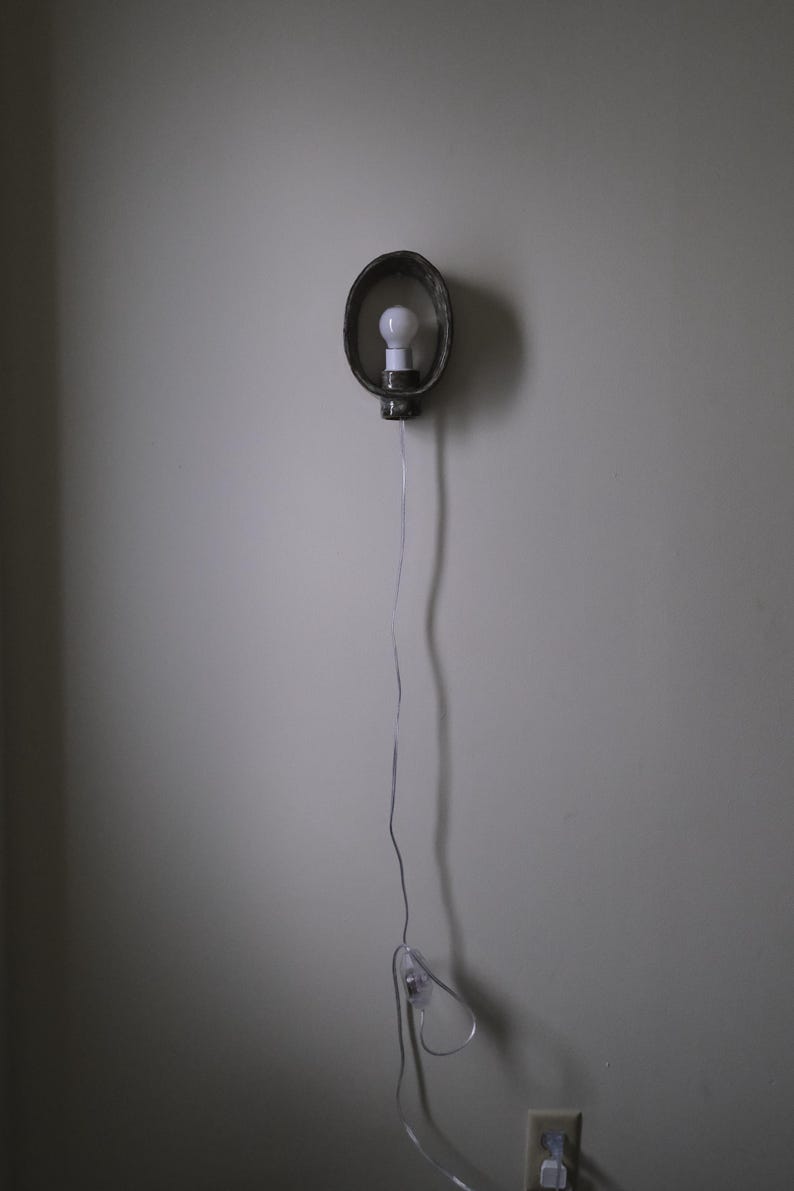 May include: A wall-mounted light fixture with a bulb inside a dark, oval-shaped metal frame. The fixture is connected to a power outlet via a thin, silver cord. The light is turned on.