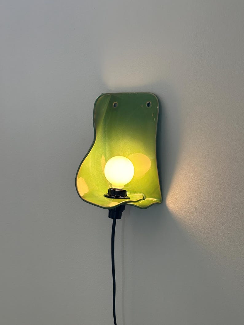Pu&ograve; includere: Una lampada da parete con un paralume in ceramica verde lime lucido, dalla forma organica e dalla silhouette curva e fluida. Una singola lampadina rotonda a luce calda &egrave; centrata nel paralume, collegata a un portalampada e a un cavo neri.