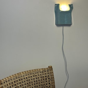 May include: A wall-mounted ceramic sconce with a speckled blue-green glaze and a U-shaped top opening. The fixture emits a warm, glowing light from within and features a thin gray power cord extending downward from the base of the ceramic unit.