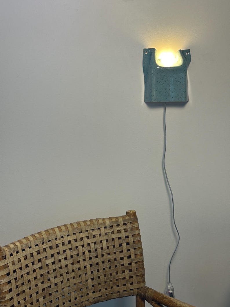 May include: A wall-mounted ceramic sconce with a speckled blue-green glaze and a U-shaped top opening. The fixture emits a warm, glowing light from within and features a thin gray power cord extending downward from the base of the ceramic unit.