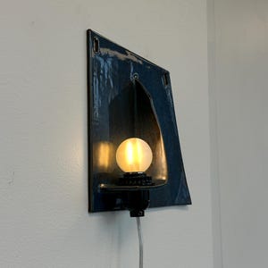 May include: A unique, wall-mounted light fixture with a dark blue, angular ceramic shade. The fixture features a warm, glowing bulb and a black base. The cord is visible, and the fixture is mounted on a white wall.