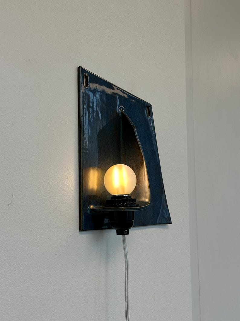 May include: A unique, wall-mounted light fixture with a dark blue, angular ceramic shade. The fixture features a warm, glowing bulb and a black base. The cord is visible, and the fixture is mounted on a white wall.