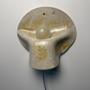 May include: A wall-mounted ceramic light fixture with a curved, organic shape and a mottled cream and yellow glaze. A single spherical white glass bulb is centered in the middle of the piece, and a thin black electrical cord hangs from the bottom.