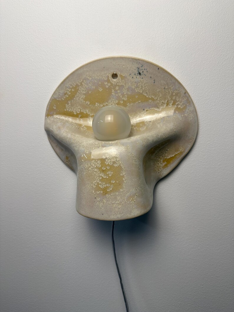 May include: A wall-mounted ceramic light fixture with a curved, organic shape and a mottled cream and yellow glaze. A single spherical white glass bulb is centered in the middle of the piece, and a thin black electrical cord hangs from the bottom.