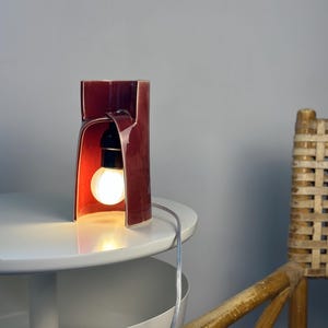 May include: A cylindrical ceramic table lamp with a deep burgundy glossy glaze. The lamp features a large arched cutout on one side, revealing a black socket and a glowing round light bulb. A thin, clear electrical cord extends from the lamp.