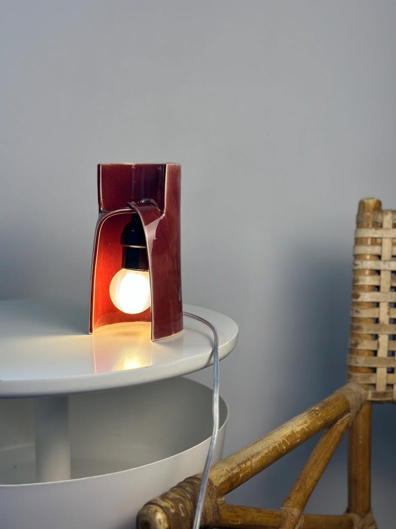 May include: A cylindrical ceramic table lamp with a deep burgundy glossy glaze. The lamp features a large arched cutout on one side, revealing a black socket and a glowing round light bulb. A thin, clear electrical cord extends from the lamp.