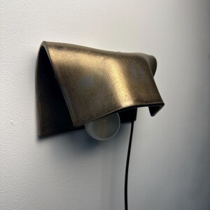 Pode incluir: Uma lumin&aacute;ria de parede com uma c&uacute;pula de metal curva em tom de bronze com acabamento fosco e texturizado. A pe&ccedil;a inclui uma l&acirc;mpada redonda branca posicionada sob a c&uacute;pula e um cabo de alimenta&ccedil;&atilde;o preto fino que se estende para baixo.