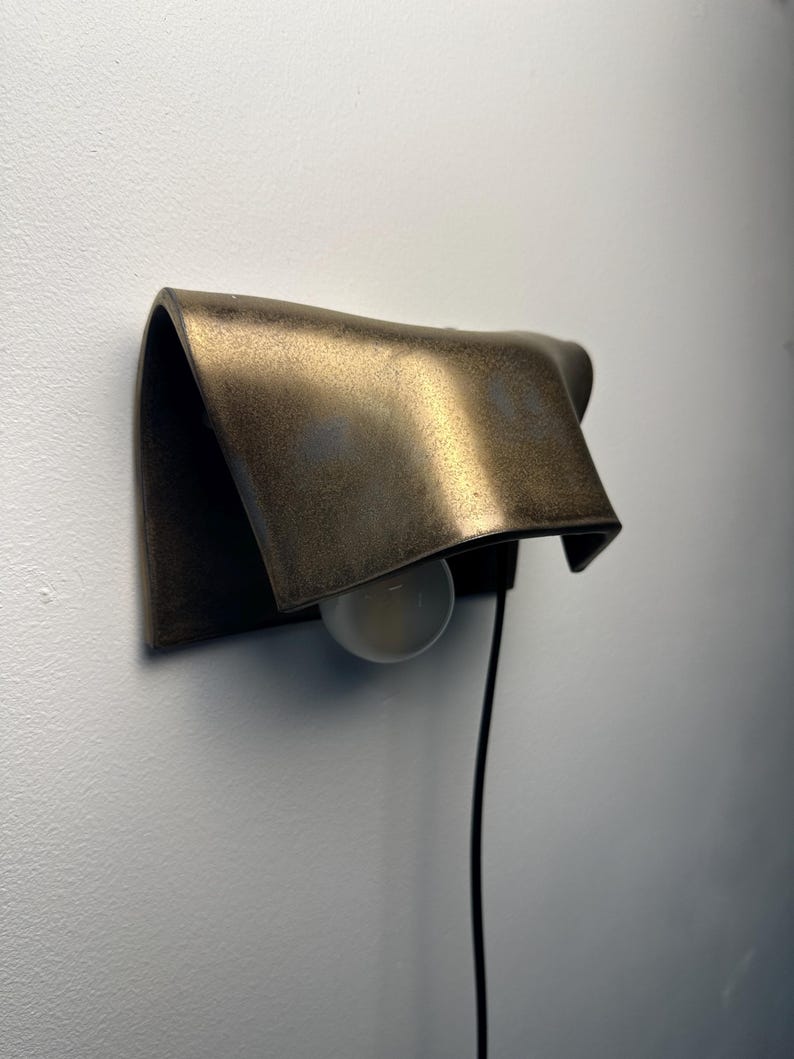 Pode incluir: Uma lumin&aacute;ria de parede com uma c&uacute;pula de metal curva em tom de bronze com acabamento fosco e texturizado. A pe&ccedil;a inclui uma l&acirc;mpada redonda branca posicionada sob a c&uacute;pula e um cabo de alimenta&ccedil;&atilde;o preto fino que se estende para baixo.