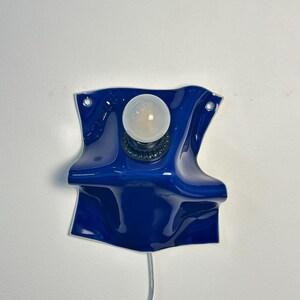 May include: A wall-mounted lamp featuring a glossy, deep blue ceramic base with a wavy, folded shape. The fixture includes a central black socket holding a single round, frosted white light bulb and a thin power cord extending from the bottom.