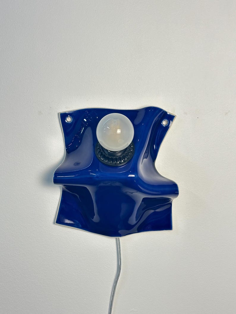 May include: A wall-mounted lamp featuring a glossy, deep blue ceramic base with a wavy, folded shape. The fixture includes a central black socket holding a single round, frosted white light bulb and a thin power cord extending from the bottom.