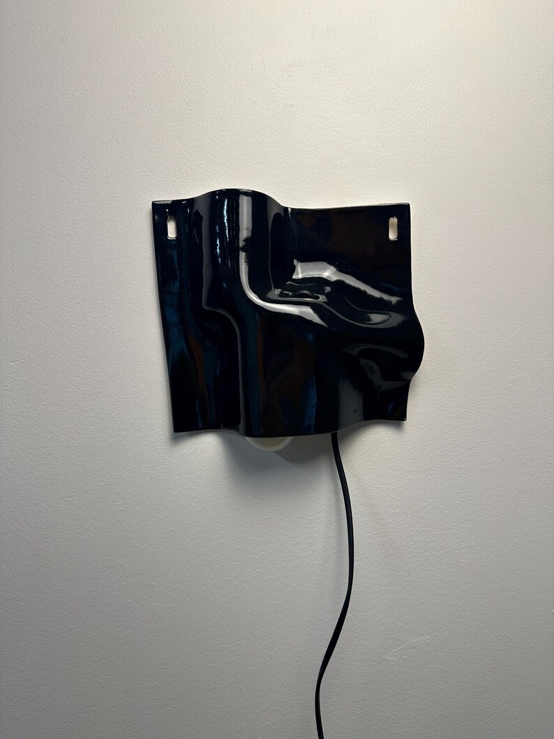 Puede incluir: Un aplique de pared negro brillante con una superficie ondulada. La pieza rectangular presenta dos peque&ntilde;os recortes rectangulares en la parte superior para el montaje y un cable de alimentaci&oacute;n negro que sale del centro inferior de la unidad.
