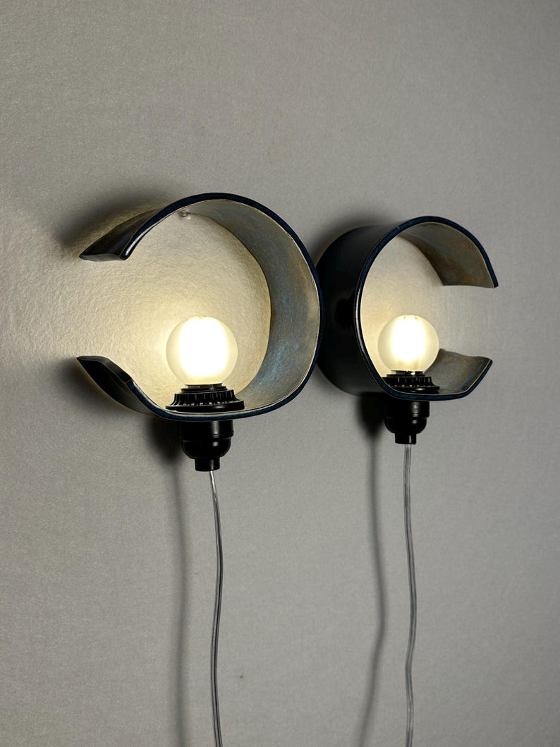 May include: Two wall-mounted lamps featuring dark, curved, C-shaped ceramic shades. Each lamp has a central, glowing spherical light bulb held by a black socket, with a thin, transparent electrical cord extending downward from the base of each fixture.