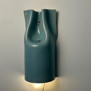 May include: A wall-mounted light fixture featuring a glossy, light blue ceramic body with a smooth, curved, and elongated silhouette. The fixture has a central opening for mounting and emits a warm, soft glow from a bulb positioned at the base.