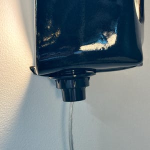 May include: A dark navy blue, glossy, rectangular wall-mounted liquid dispenser featuring a black circular nozzle at the bottom. A thin, clear, flexible plastic tube extends downward from the center of the nozzle, facilitating the flow of liquid.