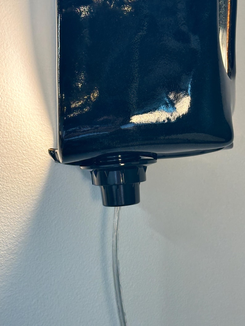 May include: A dark navy blue, glossy, rectangular wall-mounted liquid dispenser featuring a black circular nozzle at the bottom. A thin, clear, flexible plastic tube extends downward from the center of the nozzle, facilitating the flow of liquid.