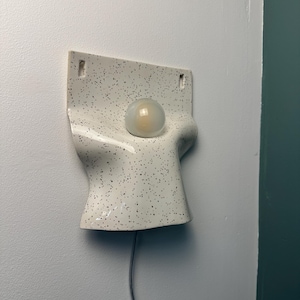 Może przedstawiać: Ścienna lampa ceramiczna z białym, nakrapianym wykończeniem i zakrzywionym, organicznym kształtem. Oprawa posiada centralny okrągły otw&oacute;r z matową żar&oacute;wką oraz dwa małe prostokątne otwory w g&oacute;rnej części służące do montażu na ścianie.