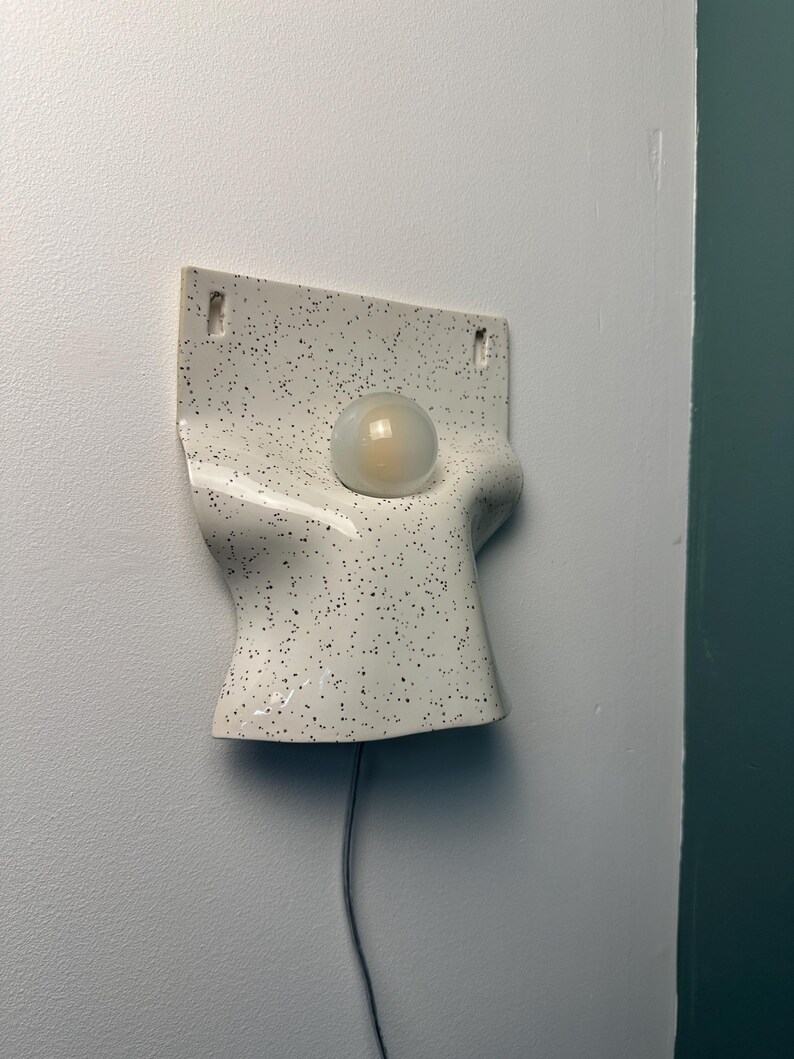 Może przedstawiać: Ścienna lampa ceramiczna z białym, nakrapianym wykończeniem i zakrzywionym, organicznym kształtem. Oprawa posiada centralny okrągły otw&oacute;r z matową żar&oacute;wką oraz dwa małe prostokątne otwory w g&oacute;rnej części służące do montażu na ścianie.