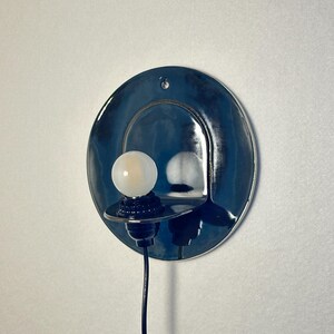 May include: A circular wall sconce featuring a glossy, deep blue ceramic backplate with a small mounting hole at the top. A black socket extends from the center, holding a small, round, frosted white light bulb. A black power cord hangs downward.
