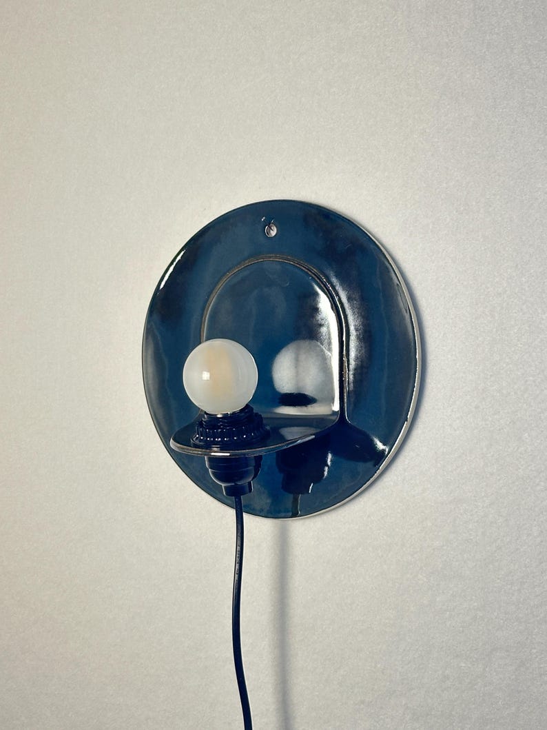 May include: A circular wall sconce featuring a glossy, deep blue ceramic backplate with a small mounting hole at the top. A black socket extends from the center, holding a small, round, frosted white light bulb. A black power cord hangs downward.