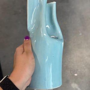 May include: A light blue ceramic vase with an irregular, organic silhouette featuring two distinct vertical openings at the top. The surface has a smooth, glossy finish with subtle variations in color tone and a wide, open base structure.