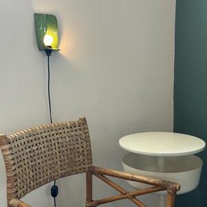 Pu&ograve; includere: Una lampada da parete con un paralume curvo in ceramica verde lucida che emette una luce calda. Sotto di essa si trova una sedia in rattan intrecciato con struttura in legno e un tavolino circolare bianco a due livelli con superficie liscia.