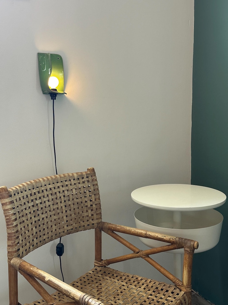 Pu&ograve; includere: Una lampada da parete con un paralume curvo in ceramica verde lucida che emette una luce calda. Sotto di essa si trova una sedia in rattan intrecciato con struttura in legno e un tavolino circolare bianco a due livelli con superficie liscia.