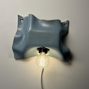 May include: A wall-mounted light fixture featuring a wavy, blue-grey ceramic shade with a smooth, matte finish. A black socket holds a single, clear, round Edison-style light bulb with a visible glowing filament, connected to a thin, light-colored cord.
