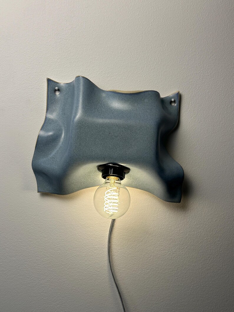 May include: A wall-mounted light fixture featuring a wavy, blue-grey ceramic shade with a smooth, matte finish. A black socket holds a single, clear, round Edison-style light bulb with a visible glowing filament, connected to a thin, light-colored cord.