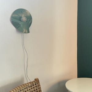 May include: A sage green ceramic wall sconce with a circular, slightly concave shape and a central white light bulb. The fixture features a downward-curving spout-like protrusion and is connected to a thin, white electrical cord hanging vertically.