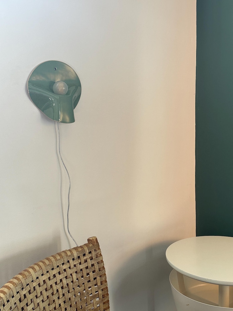 May include: A sage green ceramic wall sconce with a circular, slightly concave shape and a central white light bulb. The fixture features a downward-curving spout-like protrusion and is connected to a thin, white electrical cord hanging vertically.