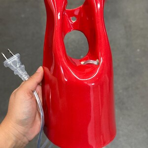 May include: A glossy red ceramic wall sconce or lamp fixture with a smooth, curved, elongated silhouette. The piece features a central circular cutout near the top, a small mounting hole, and a clear electrical cord with a standard two-prong plug.