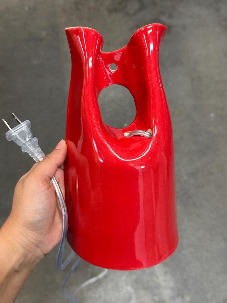 May include: A glossy red ceramic wall sconce or lamp fixture with a smooth, curved, elongated silhouette. The piece features a central circular cutout near the top, a small mounting hole, and a clear electrical cord with a standard two-prong plug.