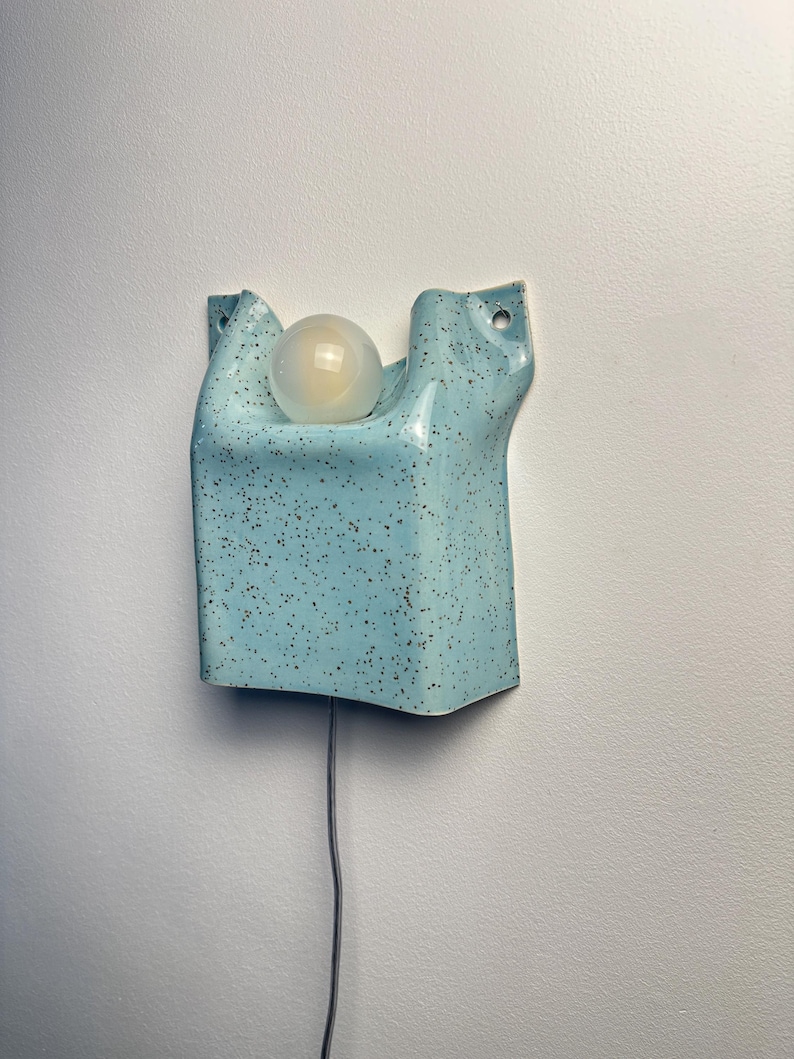 May include: A wall-mounted light fixture made of light blue ceramic with a dark brown speckled finish. The rectangular, folded-form shade features two top mounting holes and holds a single round, frosted white light bulb in the center.