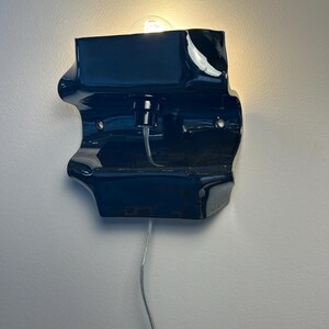 May include: A glossy, dark navy blue wall sconce with an irregular, organic shape. The fixture features a central light bulb socket, a visible clear power cord extending downward, and two metal mounting points on the front surface of the ceramic body.