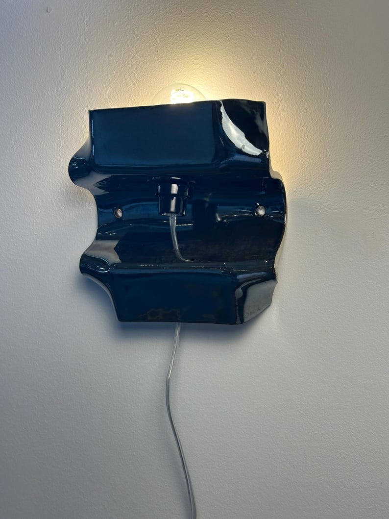 May include: A glossy, dark navy blue wall sconce with an irregular, organic shape. The fixture features a central light bulb socket, a visible clear power cord extending downward, and two metal mounting points on the front surface of the ceramic body.