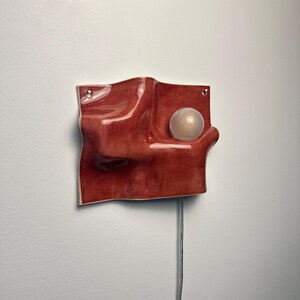May include: A wall-mounted lamp featuring a glossy, deep red, undulating ceramic form with a smooth, wave-like surface. A single, spherical, frosted white light bulb is embedded within a recessed curve of the ceramic structure, with a thin power cord.