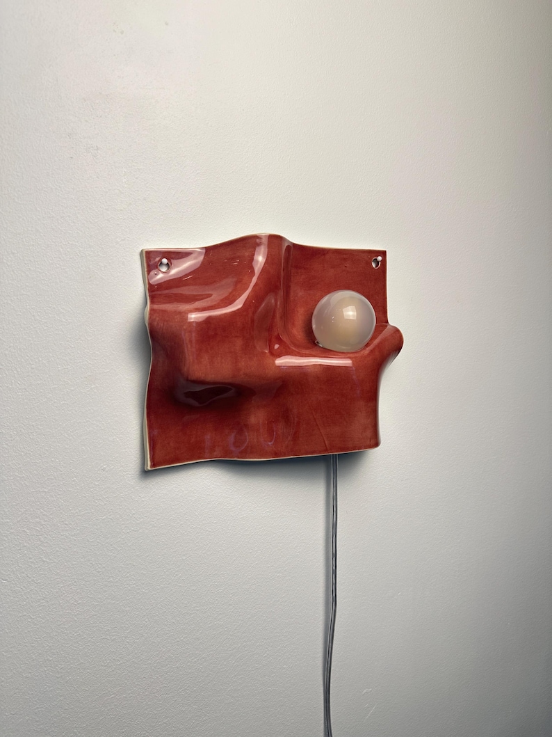 May include: A wall-mounted lamp featuring a glossy, deep red, undulating ceramic form with a smooth, wave-like surface. A single, spherical, frosted white light bulb is embedded within a recessed curve of the ceramic structure, with a thin power cord.