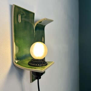 May include: A wall-mounted ceramic sconce featuring a glossy, deep green glazed finish with an organic, curved shape. The fixture includes a black socket base holding a single, glowing, frosted spherical light bulb and a black power cord.