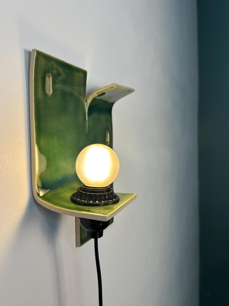 May include: A wall-mounted ceramic sconce featuring a glossy, deep green glazed finish with an organic, curved shape. The fixture includes a black socket base holding a single, glowing, frosted spherical light bulb and a black power cord.