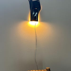 May include: A glossy black wall-mounted sconce with an irregular, organic shape. The fixture emits a warm, downward-facing glow that illuminates the wall surface below. A thin, light-colored electrical cord hangs vertically from the base of the lamp.