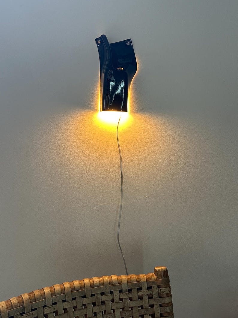 May include: A glossy black wall-mounted sconce with an irregular, organic shape. The fixture emits a warm, downward-facing glow that illuminates the wall surface below. A thin, light-colored electrical cord hangs vertically from the base of the lamp.