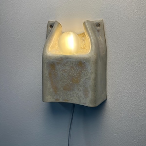 May include: A wall-mounted light fitting featuring a textured, off-white ceramic housing with a curved, open top design. A single, spherical, frosted glass bulb sits within the central recess, emitting a warm, soft glow. A thin power cord extends downwards.