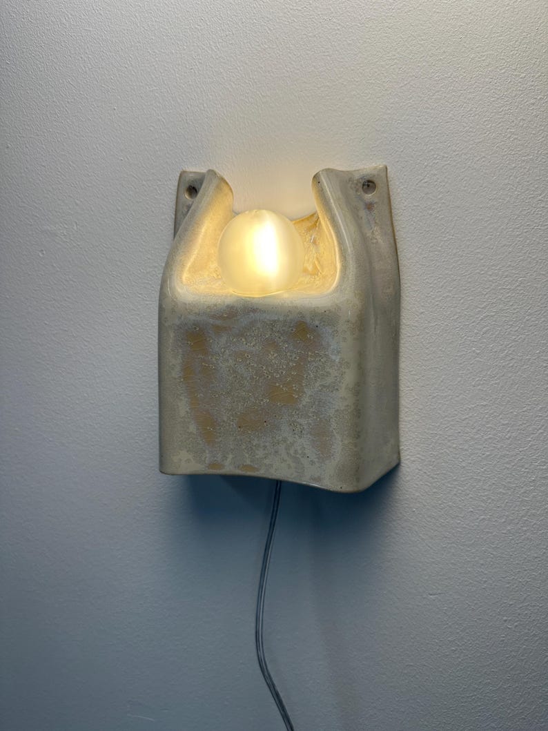 May include: A wall-mounted light fitting featuring a textured, off-white ceramic housing with a curved, open top design. A single, spherical, frosted glass bulb sits within the central recess, emitting a warm, soft glow. A thin power cord extends downwards.